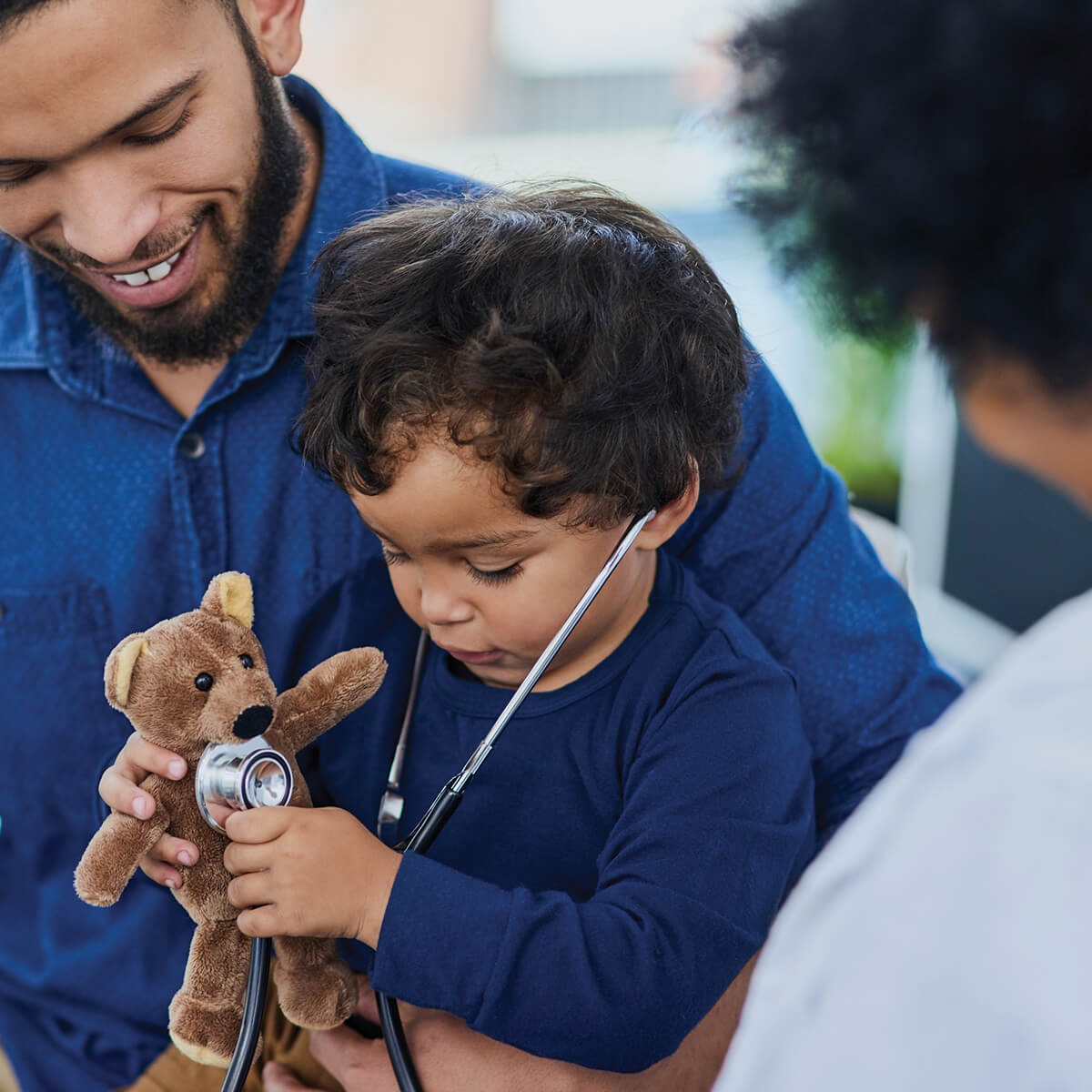 A father sitting with his son at the doctor's office, while the son tries to listen to a teddy bear's chest with a stethoscope