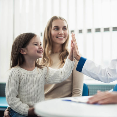 A young girl sitting in her mother's lap in the doctor's examination room, and giving the doctor a high five.