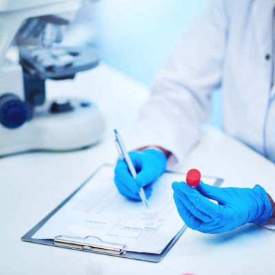 A technician in a lab filling out paperwork with their right hand, and holding a vial of blood in their right hand.